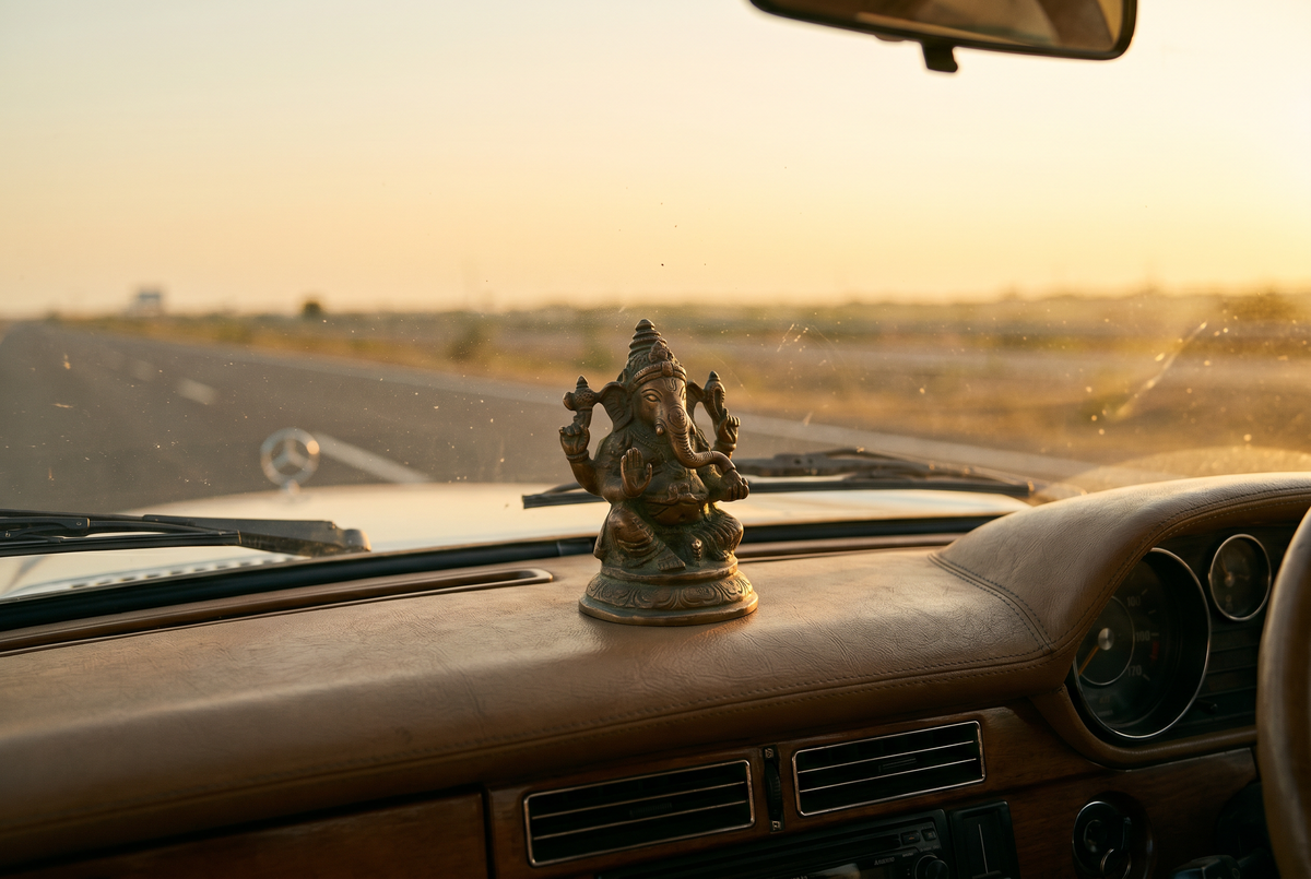 A small sculpted Indian cultural figure on a car dashboard at golden hour, the open road ahead through the windshield.