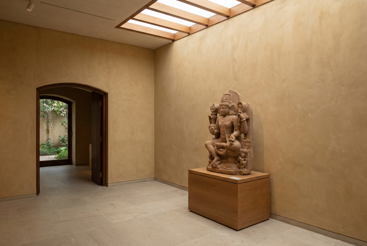 A museum-quiet gallery with warm paper-coloured walls, soft daylight from above, and a single sculpted Indian cultural figure on a central plinth.